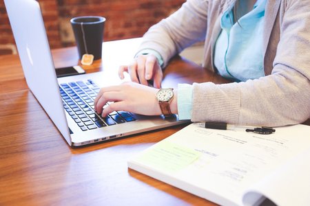 Eine Person die am Laptop arbeitet. Neben ihr eine Tasse Tee und ein offenes Buch. Gesicht ist nicht zu erkennen.