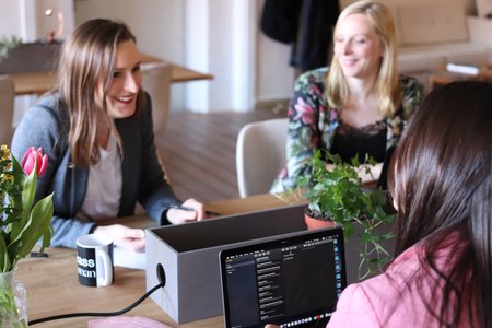 Drei Frauen beim arbeiten, in einem Meeting. Am Tisch stehen Laptops und Arbeitsmaterialien