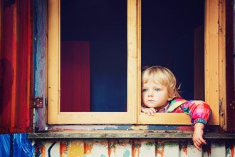 Kleiner blonder Junge sitzt an einem Fenster uns starrt in die Ferne