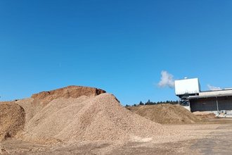 Mehrere große Haufen aus Holzschnitzeln liegen unter freiem Himmel auf einem Industrieplatz. Im Hintergrund steht ein modernes Industriegebäude mit einem weißen Aufbau, aus dem etwas Dampf aufsteigt. Der Himmel ist klar und blau.