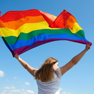 Eine junge Frau die vor blauem Himmel steht und über Kopf eine Regenbogenflagge (Prideflagge) hält.