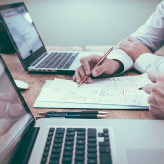 Arbeitstisch mit zwei Laptops und Papierunterlagen, Arme von zwei Personen bei der Arbeit sichtbar
