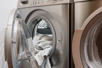 Silberne Waschmaschine und daneben gleich ein Trockner mit geöffneter Trommel, wo etwas Wäsche raushängt.